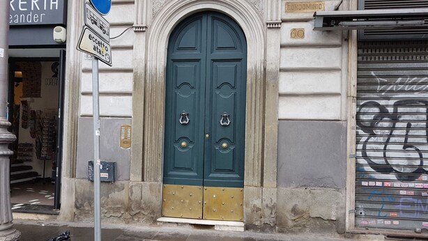 Una puerta doble verde con aldabas de latón, un letrero encima y un persiano cerrado a la derecha.