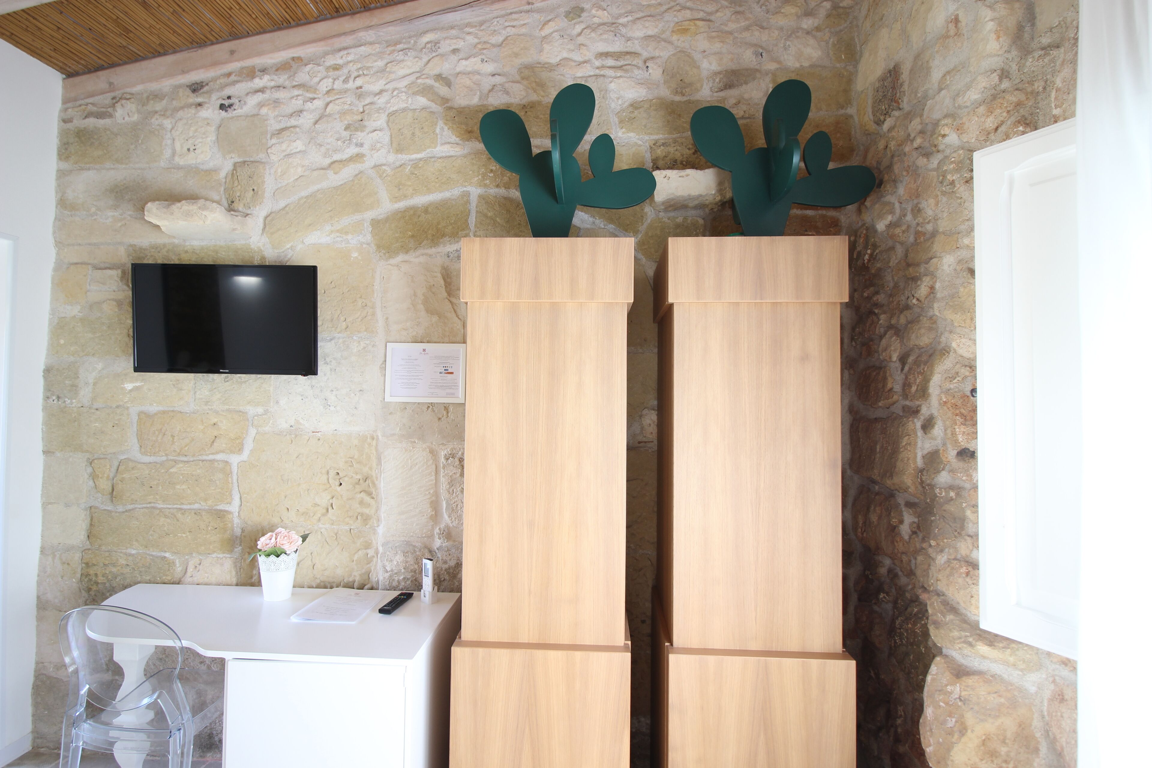 Ein Zimmer mit einer Steinwand, einem flachbildfernseher, der daran befestigt ist, einem weißen Schreibtisch mit einem Blumenstrauß darauf und zwei Holzschränken mit Kakteen-Dekorationen obenauf.