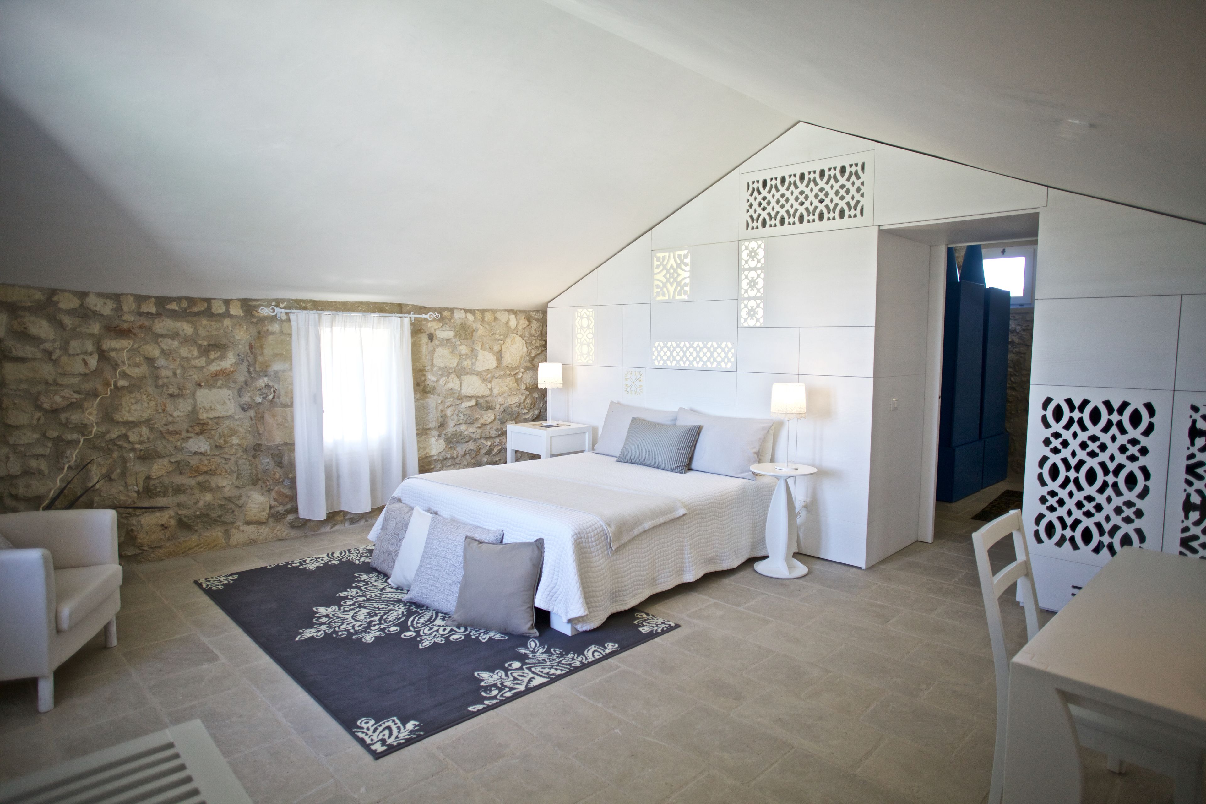 Un dormitorio con pared de piedra, una cama con ropa de cama blanca, una alfombra blanco y negro, una silla blanca, una pequeña mesa con una lámpara y un aparador azul.