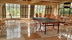 A spacious room with a ping pong table, several chairs, and large windows offering a view of greenery. - The Kingdom (Port Blair)