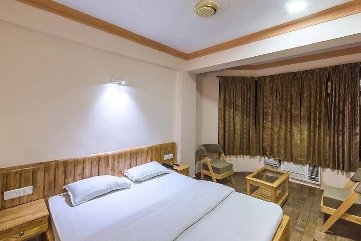 A hotel room with a wooden headboard, a bed with white linens, a bedside table, a chair, a small table, and a window with patterned curtains.