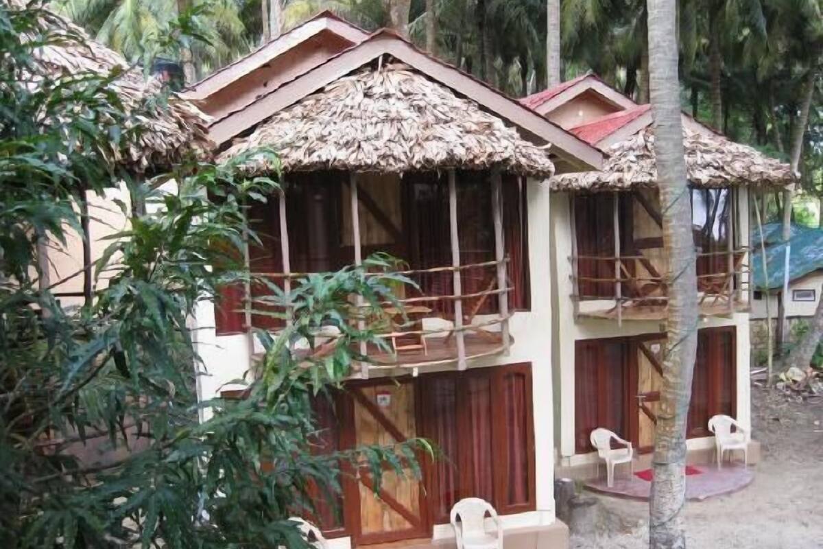A bungalow with a thatched roof, wooden balconies, and a veranda with plastic chairs.