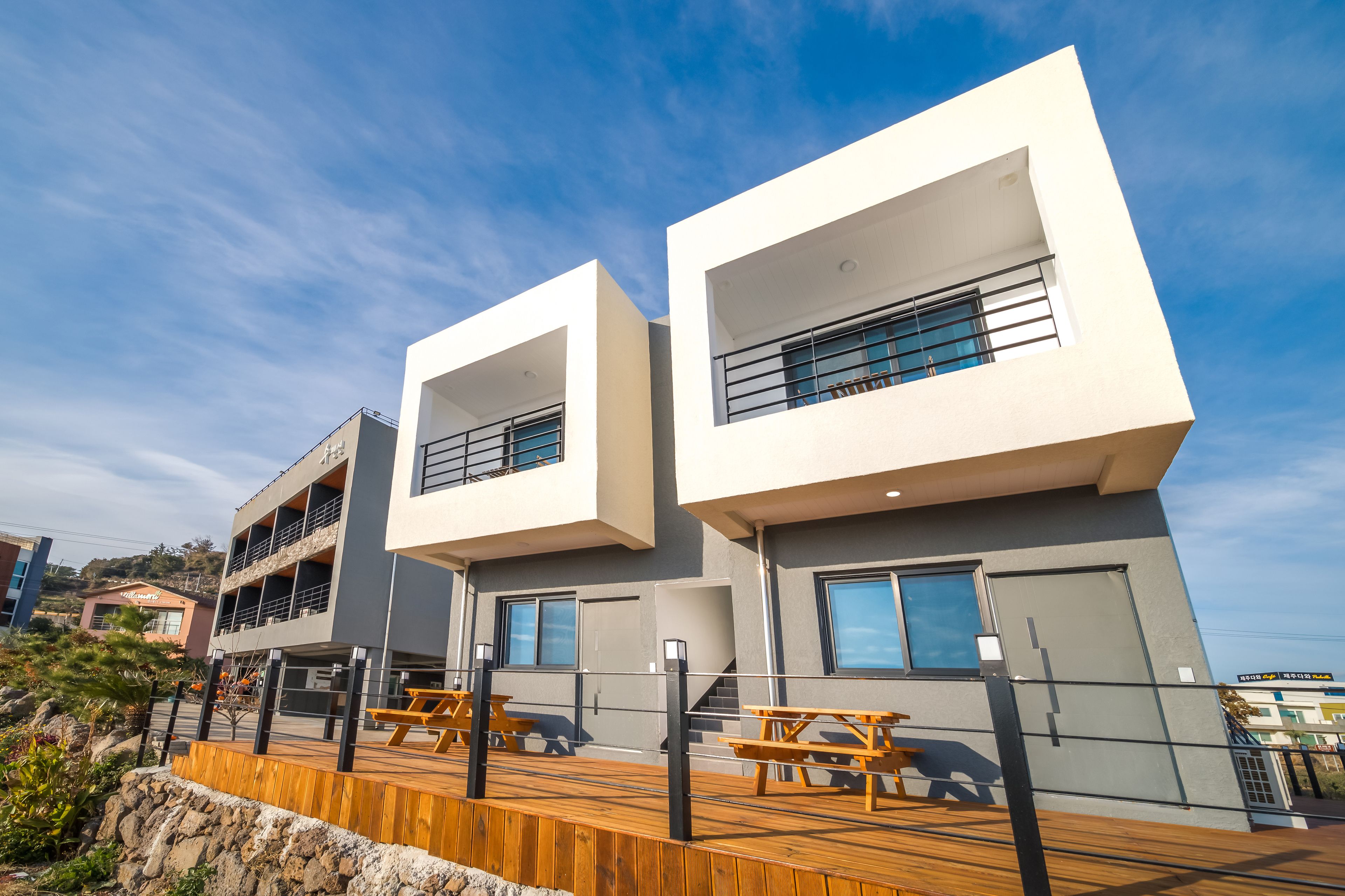 Un edificio moderno con balcones de dos pisos, terraza exterior de madera con mesas de picnic y cielo despejado.