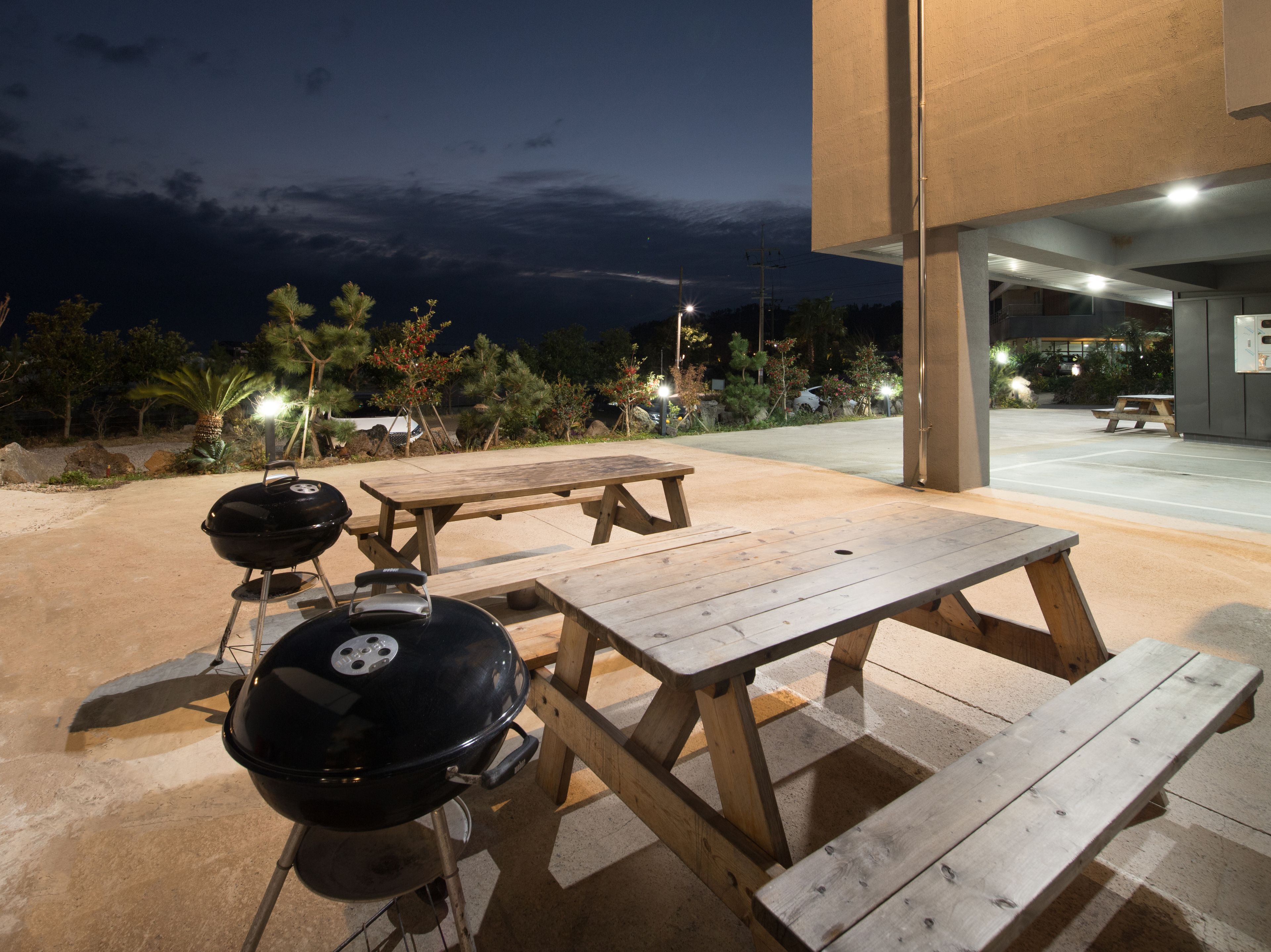 An outdoor patio area with a barbecue grill, picnic tables, and a view of a parking lot and building in the background.