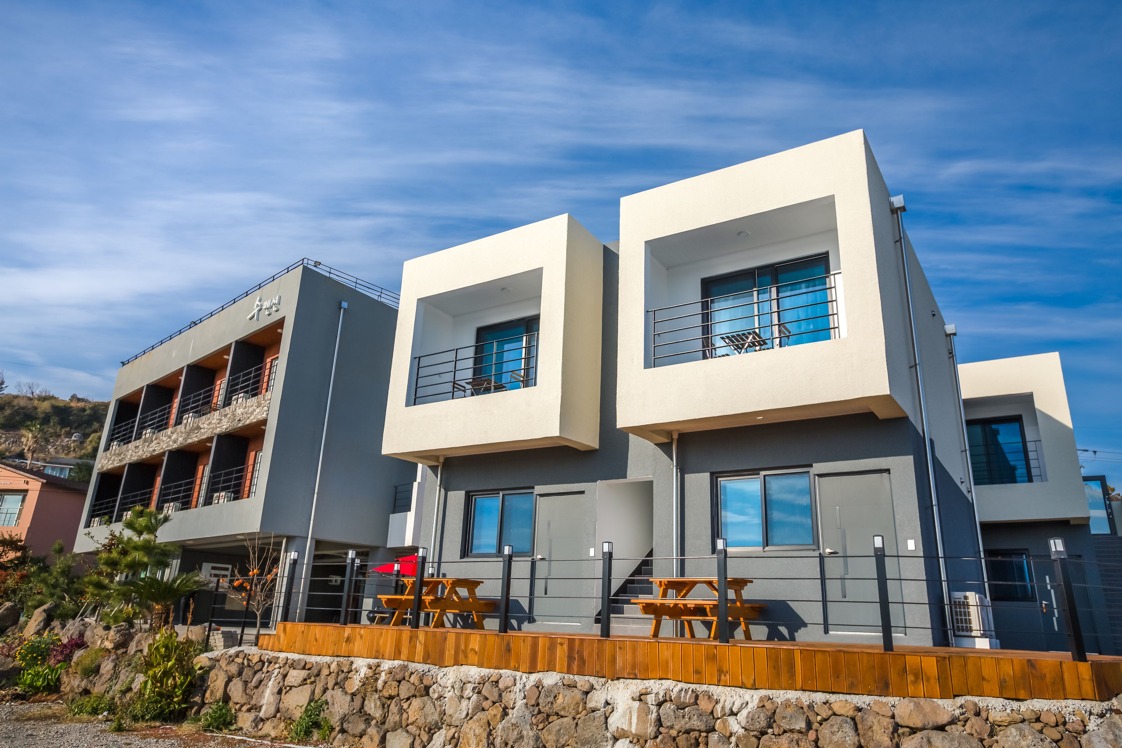 A modern two-story building with a stone retaining wall, wooden deck, and outdoor seating.