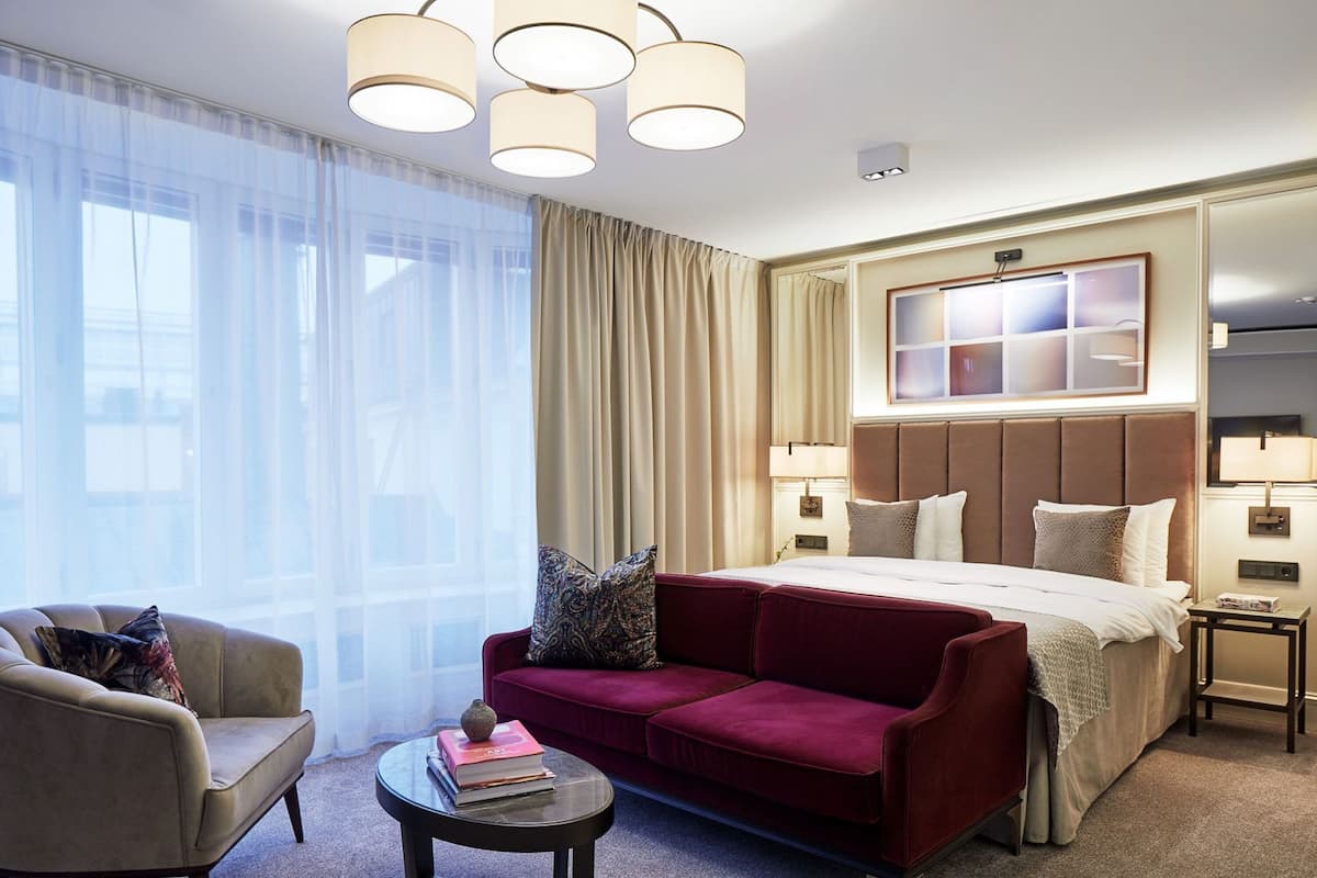 A hotel room with a bed, a red sofa, a chair, a small table with books, and a large window with sheer curtains.