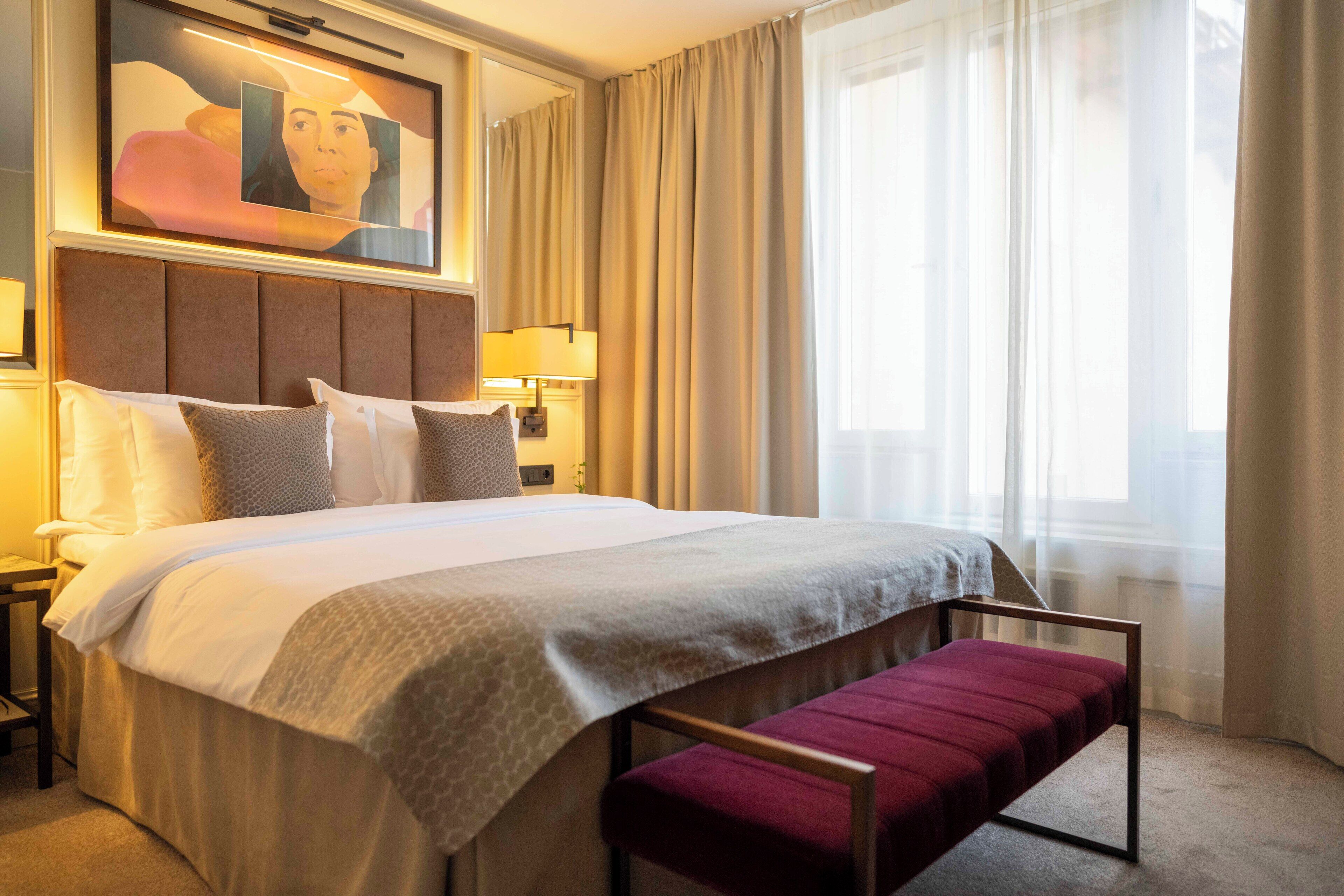 A hotel room with a large bed, a bench, a painting on the wall, and a window with curtains.