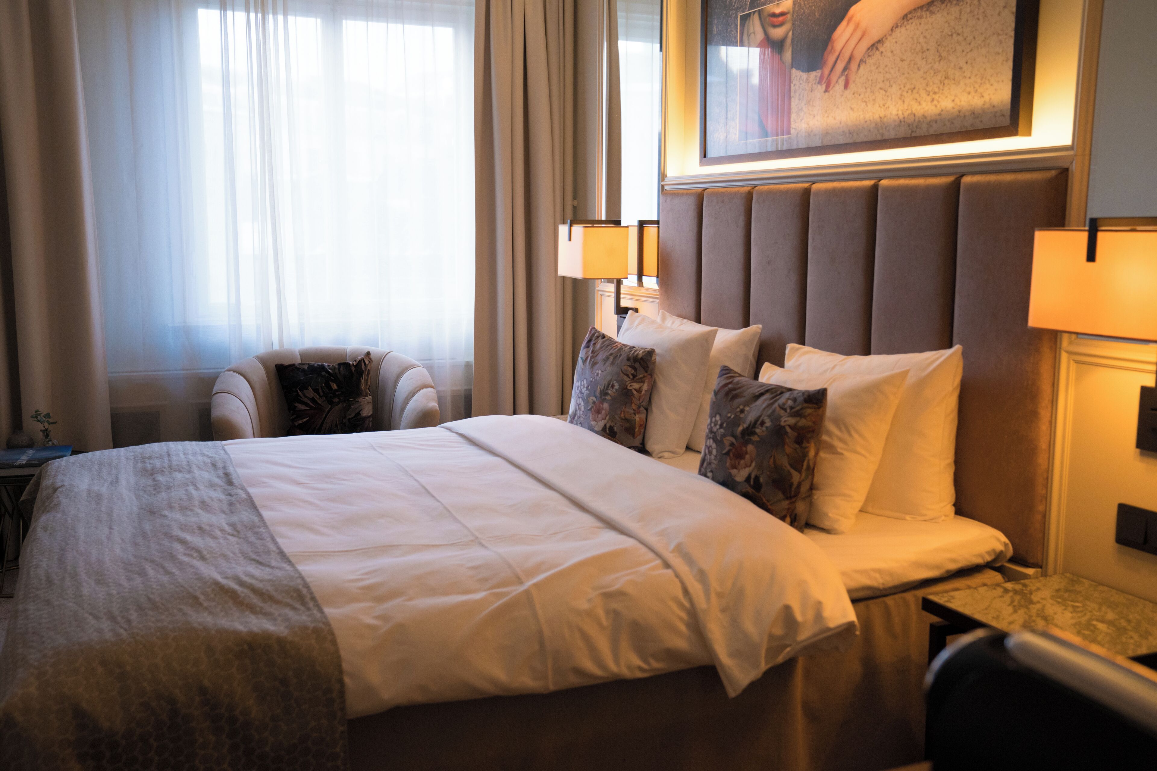 A hotel room with a large bed, two bedside lamps, a headboard, and a painting above the bed.