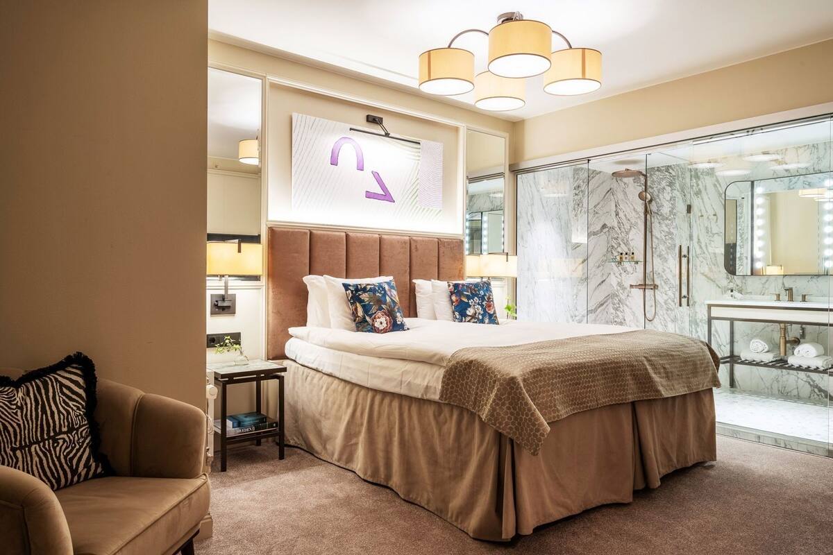 A hotel room with a large bed, a sitting area, a bathroom with a marble shower, and a large mirror with lights.