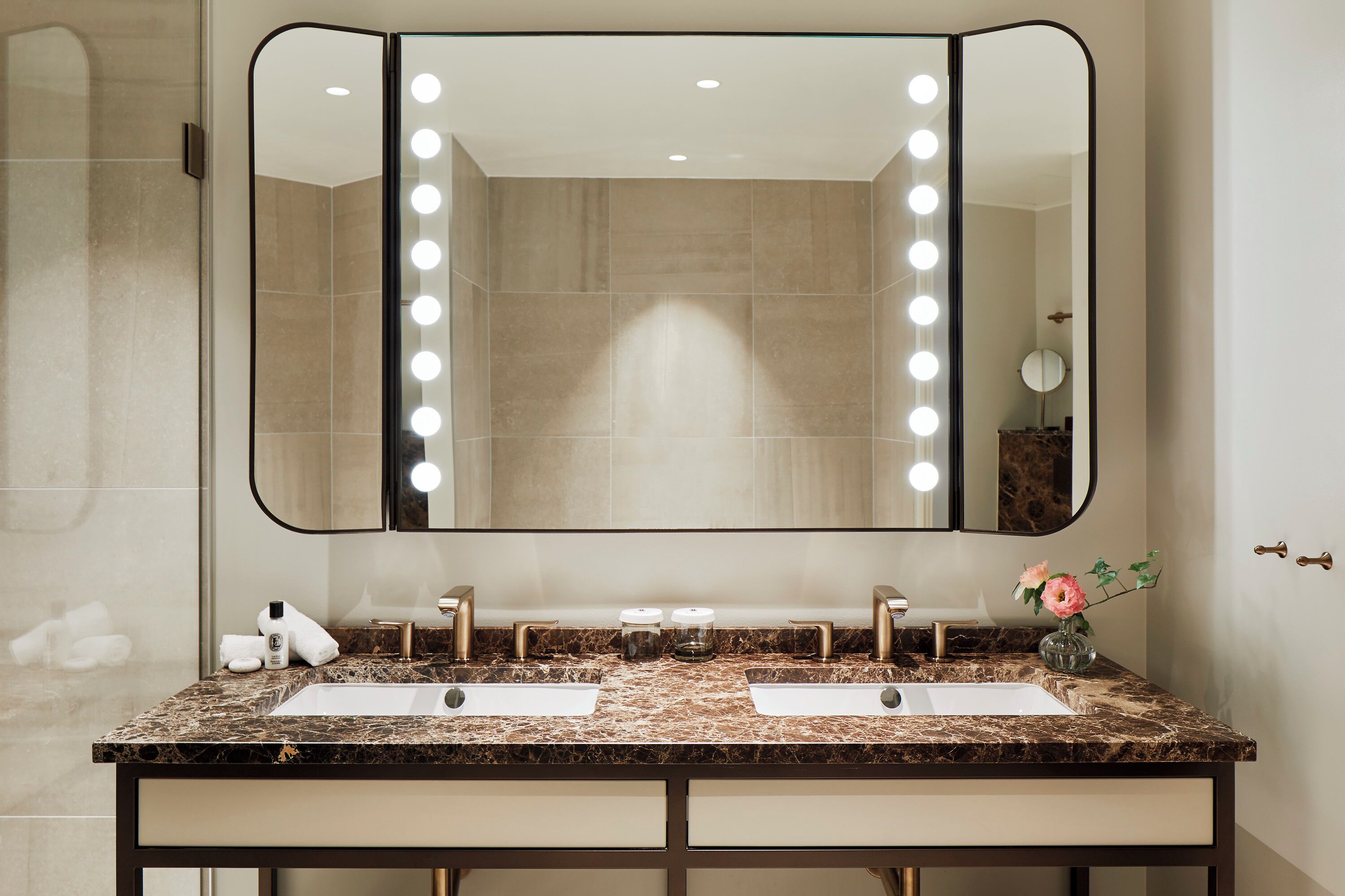 A bathroom with two sinks, a large mirror with lights, and a vase with flowers.