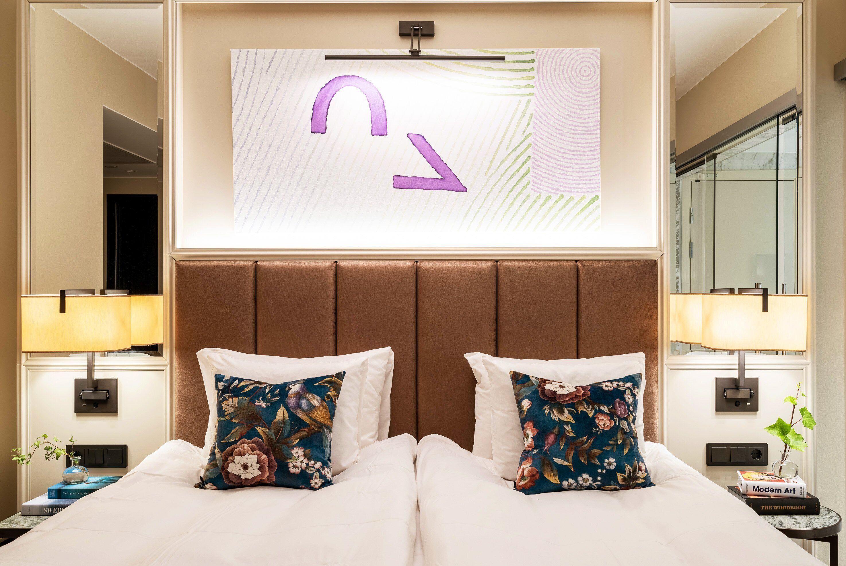 Quarto de hotel com uma cama grande, duas almofadas decorativas, uma TV na parede e uma mesa de cabeceira com abajur e um livro.
