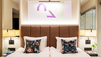 Quarto de hotel com uma cama grande, duas almofadas decorativas, uma TV na parede e uma mesa de cabeceira com abajur e um livro.