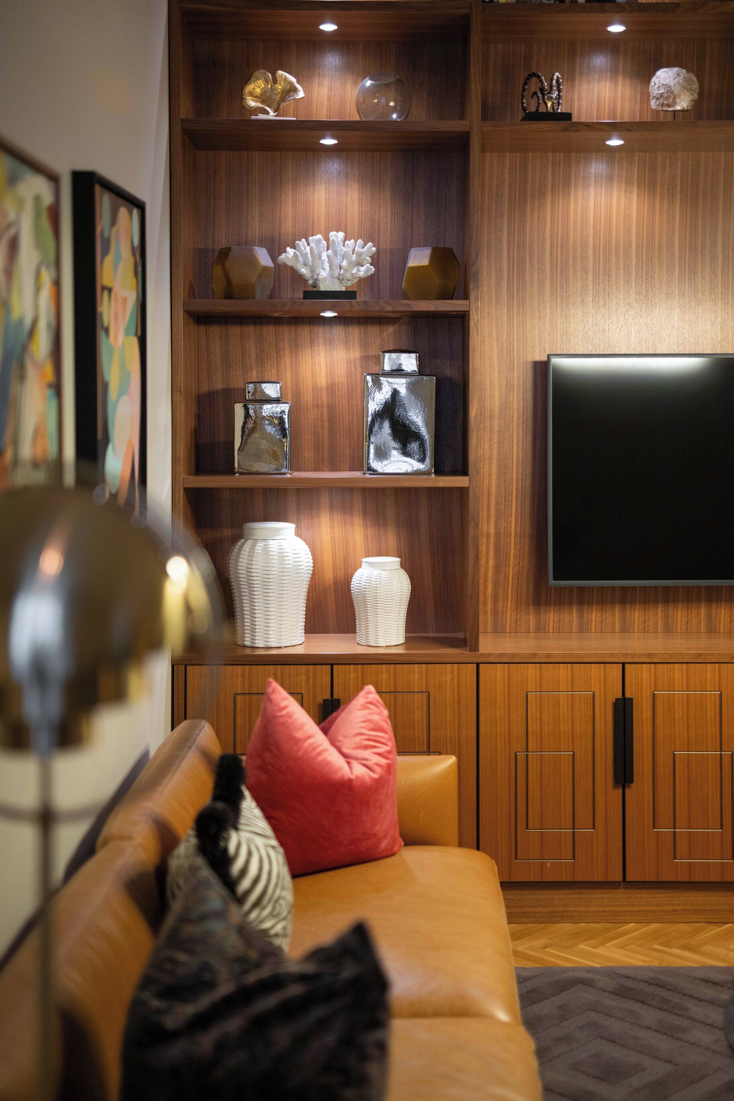 Un salon moderne avec un canapé en cuir, un téléviseur à écran plat, des étagères en bois ornées d’objets décoratifs et un parquet à motifs en chevrons.