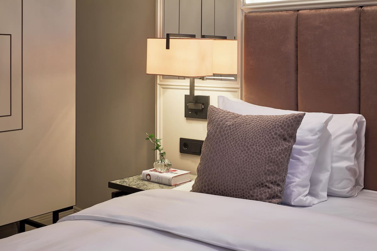 A neatly made bed with a white and brown pillow, a bedside table with a book and a vase, and a wall-mounted lamp.