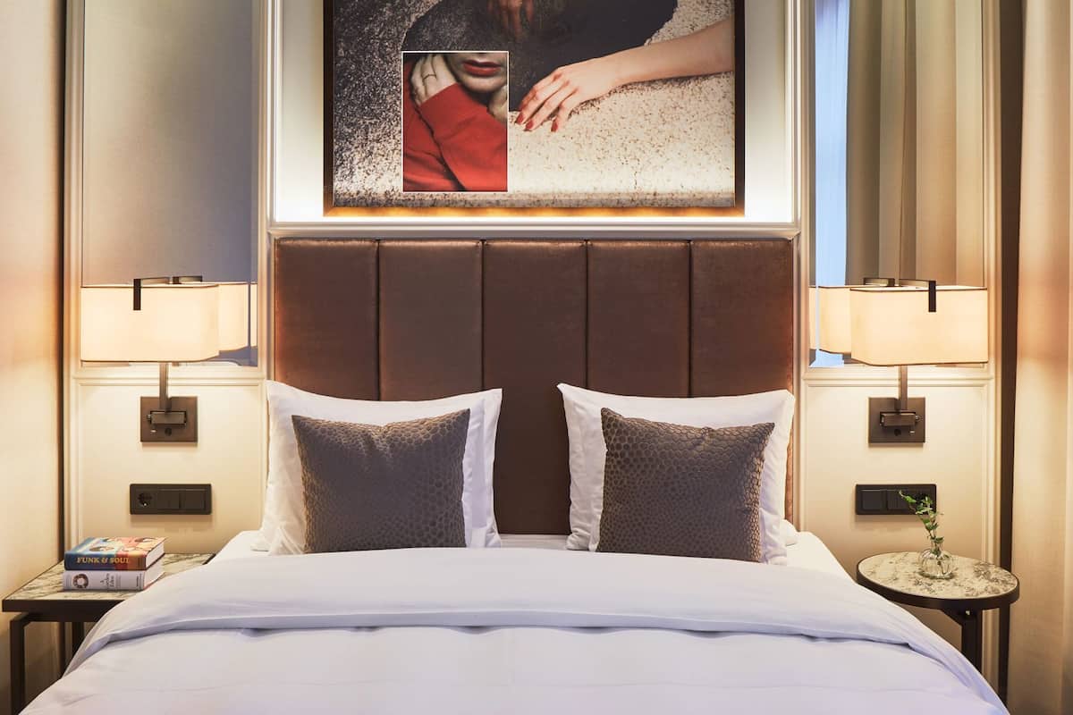 A hotel room with a large bed, two bedside lamps, a nightstand with a book, and a framed picture above the bed.