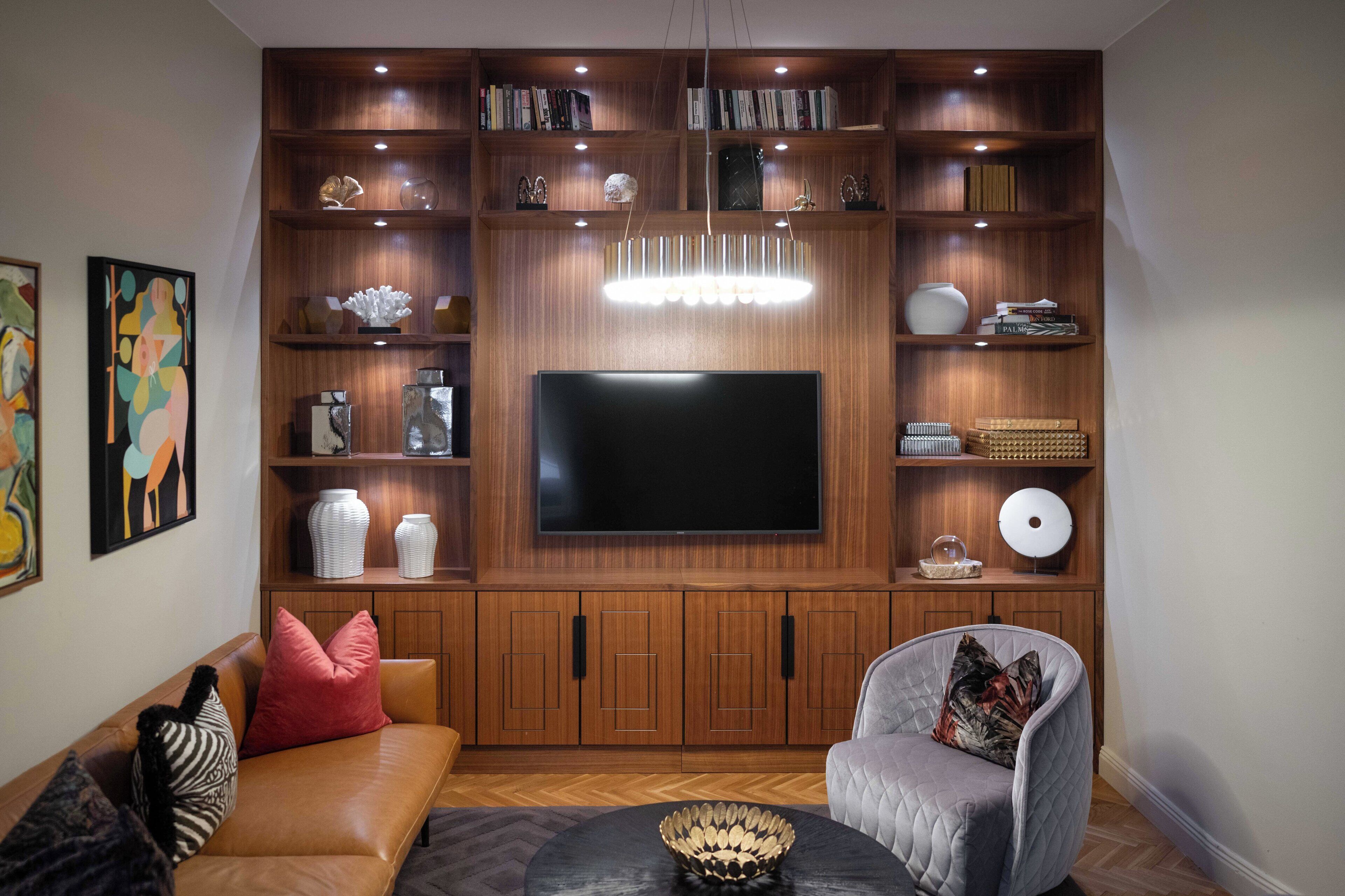 A living room with a large flat-screen TV, wooden shelving with books and decorative items, a leather sofa, and a grey armchair.