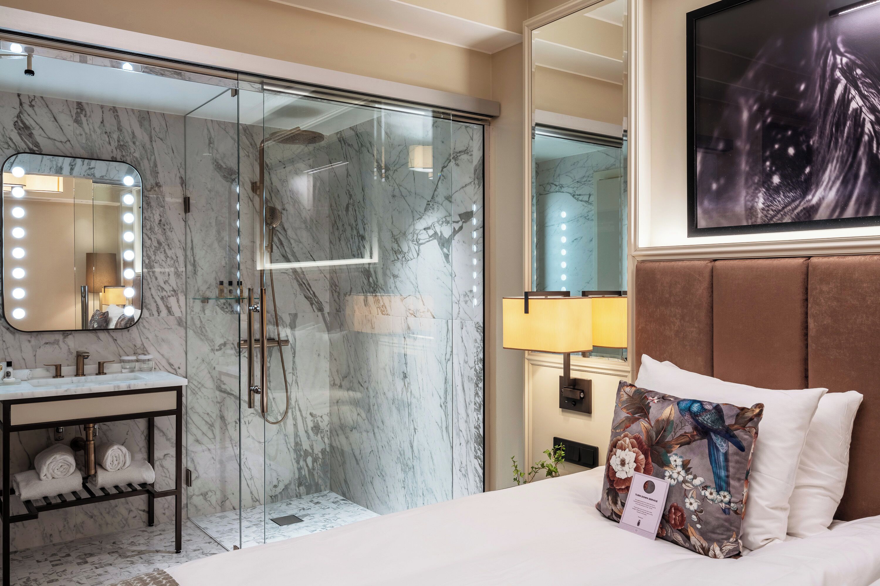 A modern hotel room with a marble bathroom, a vanity with a mirror, a bed with a decorative pillow, and a mounted artwork.