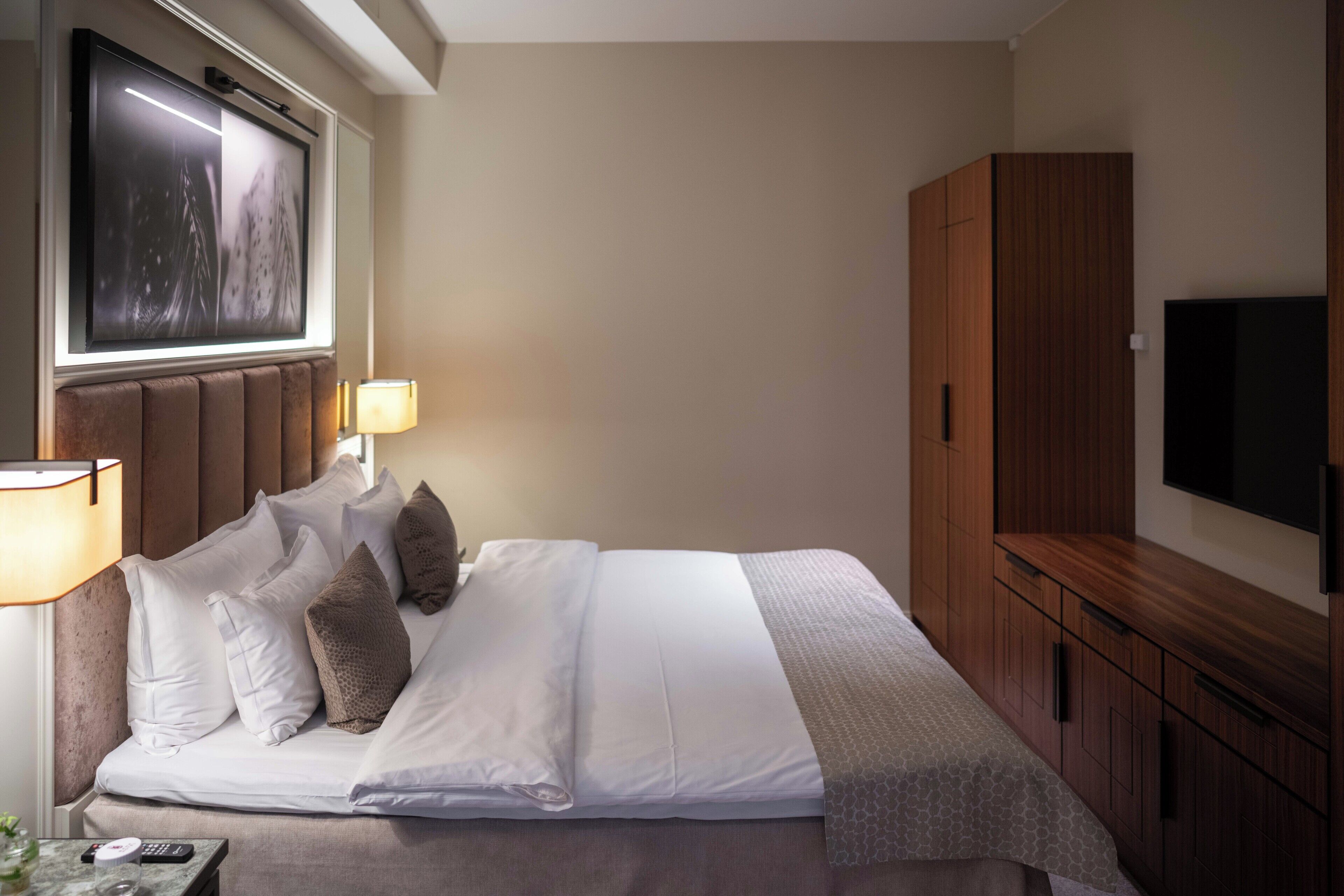A hotel room with a large bed, a TV mounted on the wall, a wooden wardrobe, and a bedside table with a lamp.