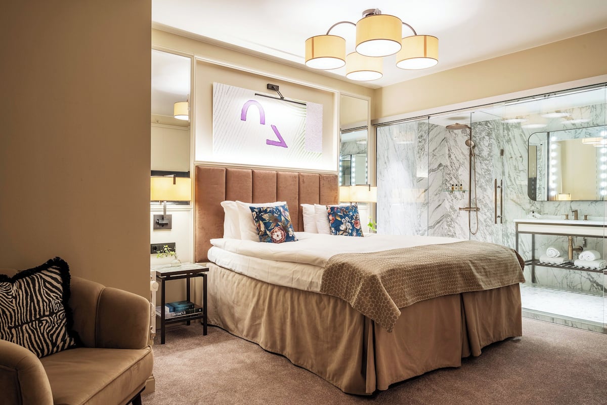 A hotel room with a large bed, a nightstand, a sofa, a bathroom with a marble shower, and a large mirror.