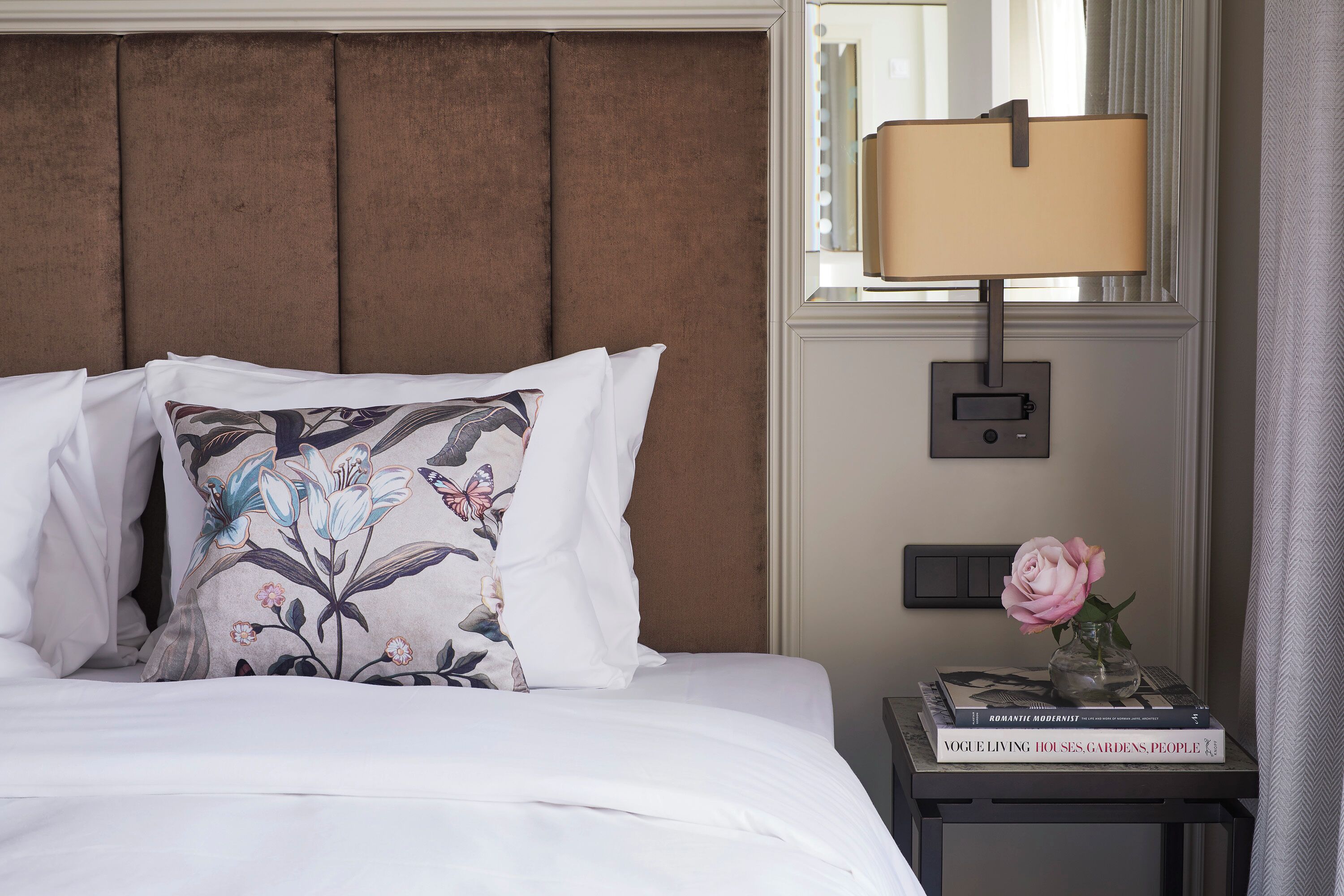 Una cama bien hecha con almohadillas florales, una mesita de noche con lámpara y un jarrón con una rosa.