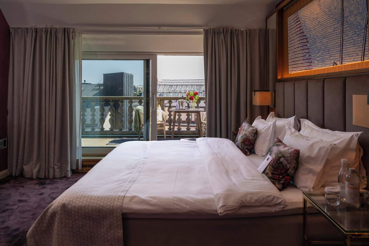 A hotel room with a large bed, a glass table with a bottle and glass, and a view of the outside balcony.