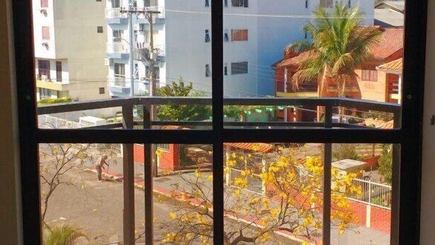 A room with a large window overlooking a residential area, featuring a balcony with a tiled floor and a metal railing. - Residencial Salinas (Florianópolis)