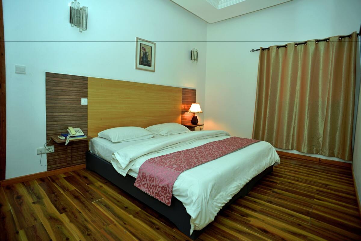 A hotel room with a wooden floor, a bed with white linens, a red patterned throw, a bedside lamp, and a telephone on the nightstand.