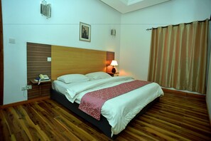 Une chambre d’hôtel avec un plancher en bois, un lit recouvert de draps blancs, un plaid à motifs rouges, une lampe de chevet et un téléphone sur la table de chevet.