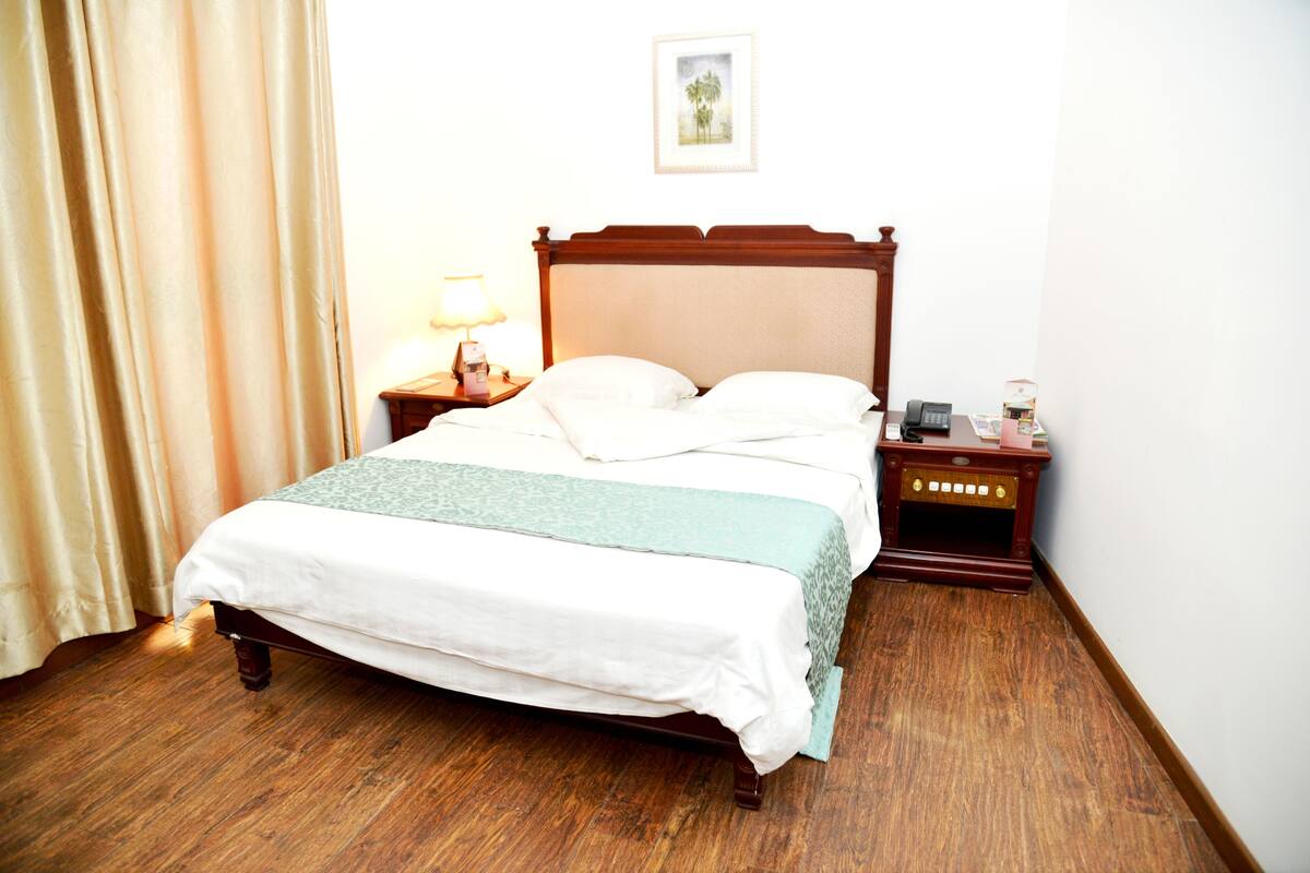 A neatly made bed with a wooden headboard, a bedside table with a lamp, and a small wooden cabinet with a telephone and a box.