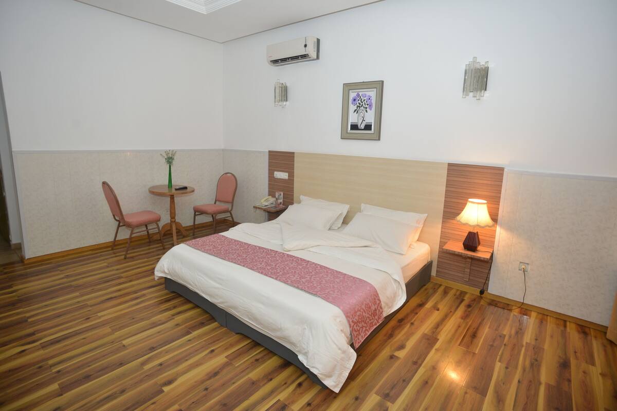 A hotel room with a wooden floor, a bed with white and pink bedding, a bedside table with a lamp, a chair, a small table with a vase, and a wall-mounted air conditioner.