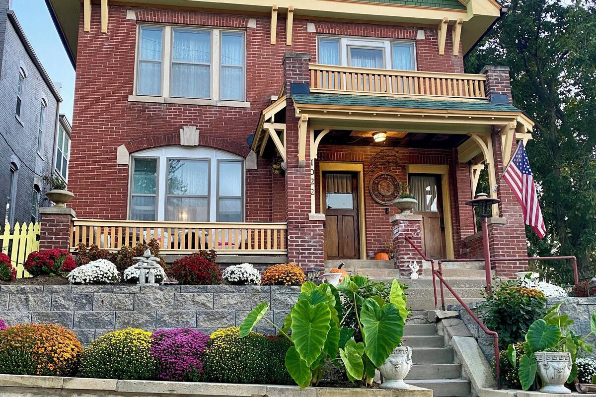 A two-story brick house with a green roof, a front porch with a railing, and a small American flag.