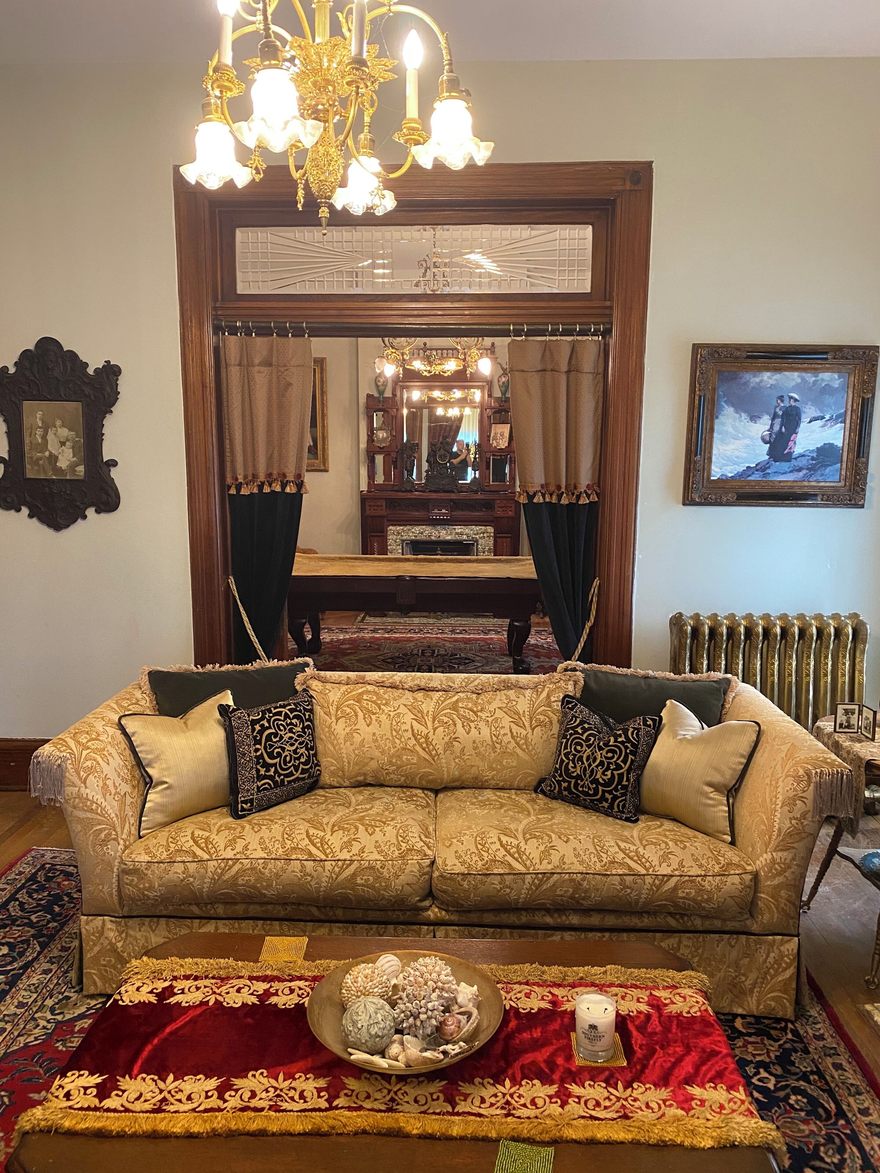 A room with a large ornate mirror, a chandelier, a sofa with patterned cushions, a decorative bowl on a red rug, and a framed picture on the wall.