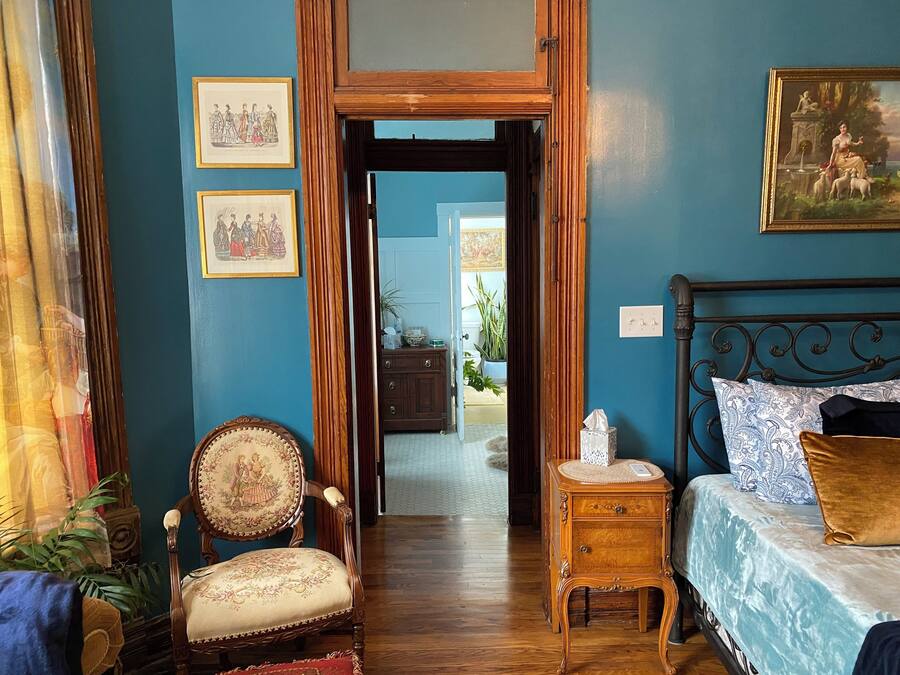 A room with a bed, a wooden chair, a small table, and a door leading to another room.