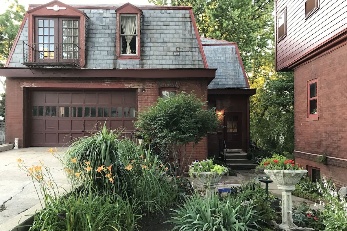 A two-story house with a garage, a front porch, and a garden with various plants and flowers.