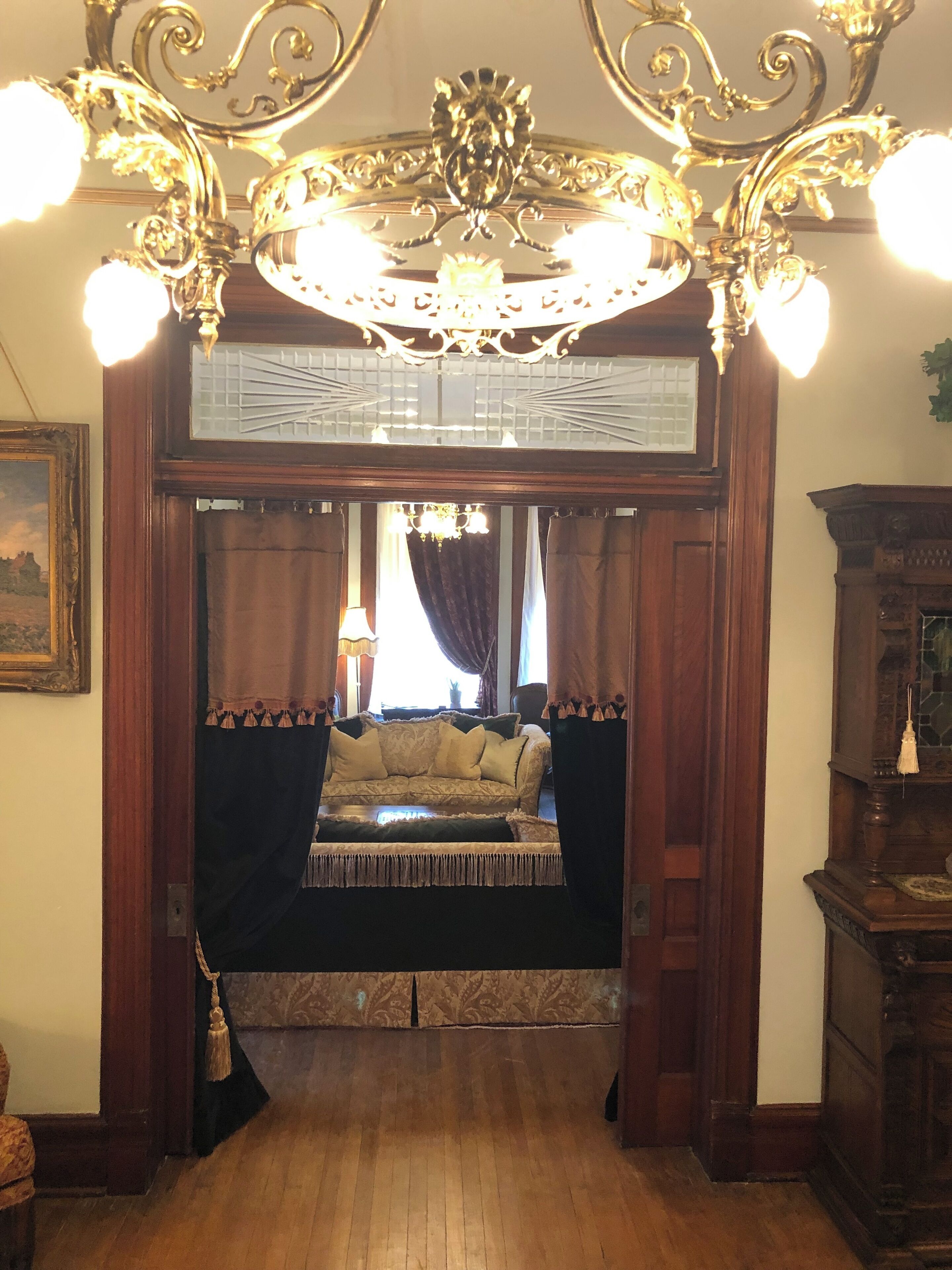 A room with a wooden floor, a sofa with a patterned cover, a chandelier, and a mirror above the sofa.