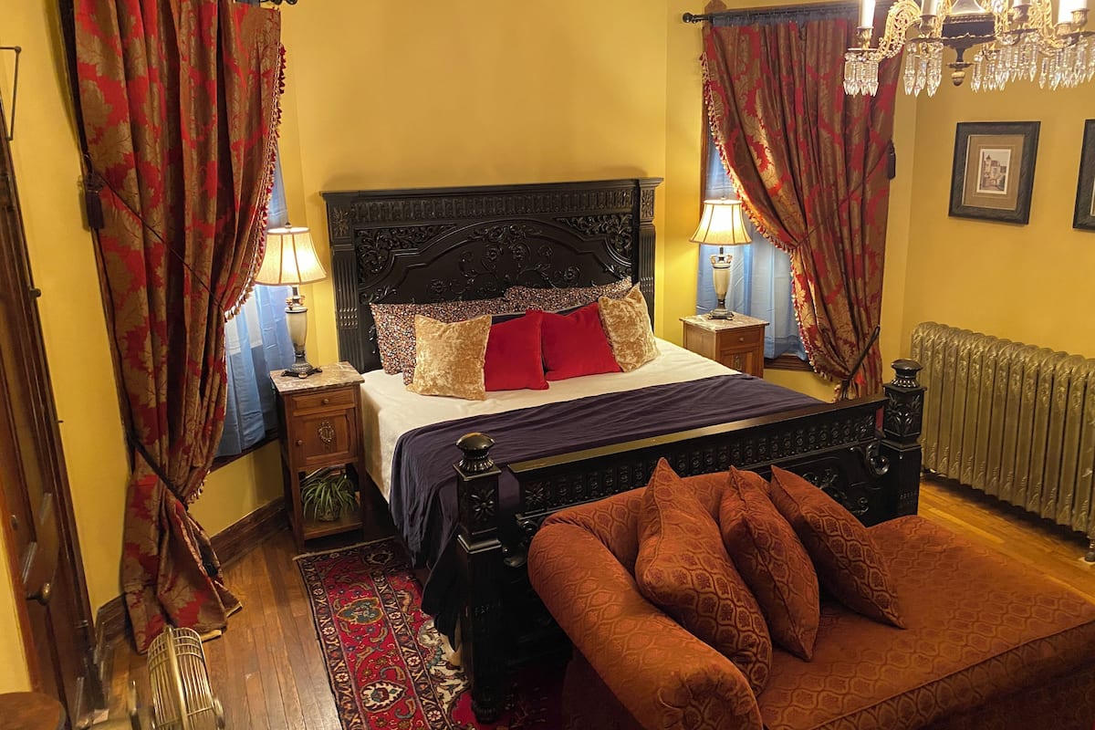 A bedroom with a four-poster bed, a wooden nightstand, a red throw pillow, a patterned rug, and a radiator.