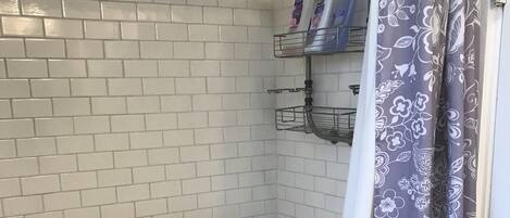 Zona de ducha con paredes de azulejos blancos, un cabezal de ducha, un organizador de ducha con artículos de aseo y una cortina de ducha con estampado floral.