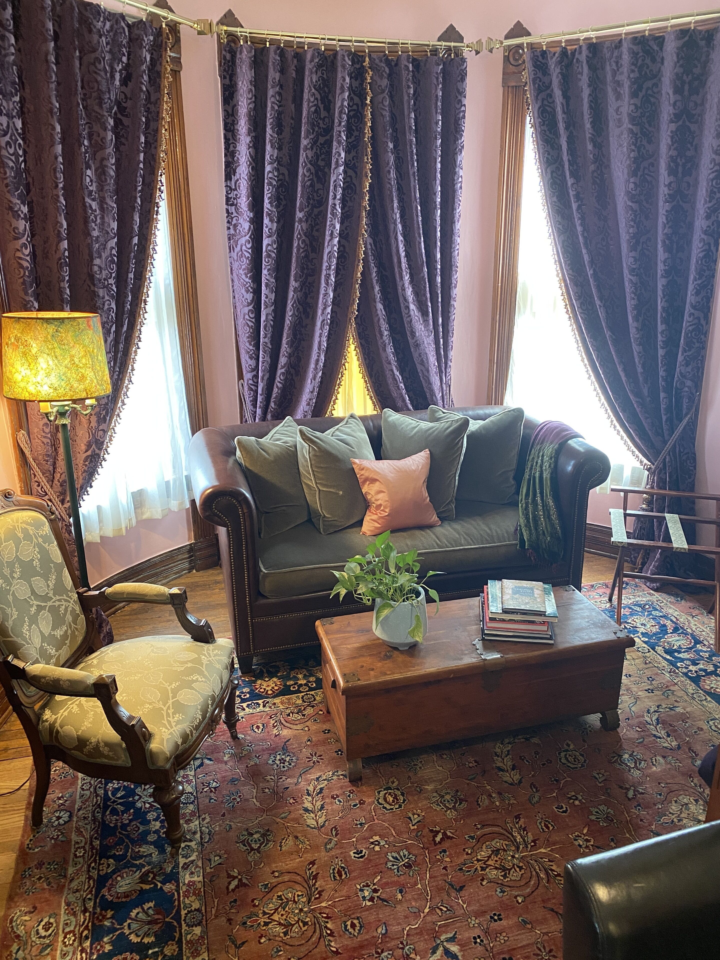 Una sala de estar con un sofá verde oscuro, cojines estampados, una mesa de centro de madera con una planta y libros, un sillón estampado y un ventanal con cortinas moradas.