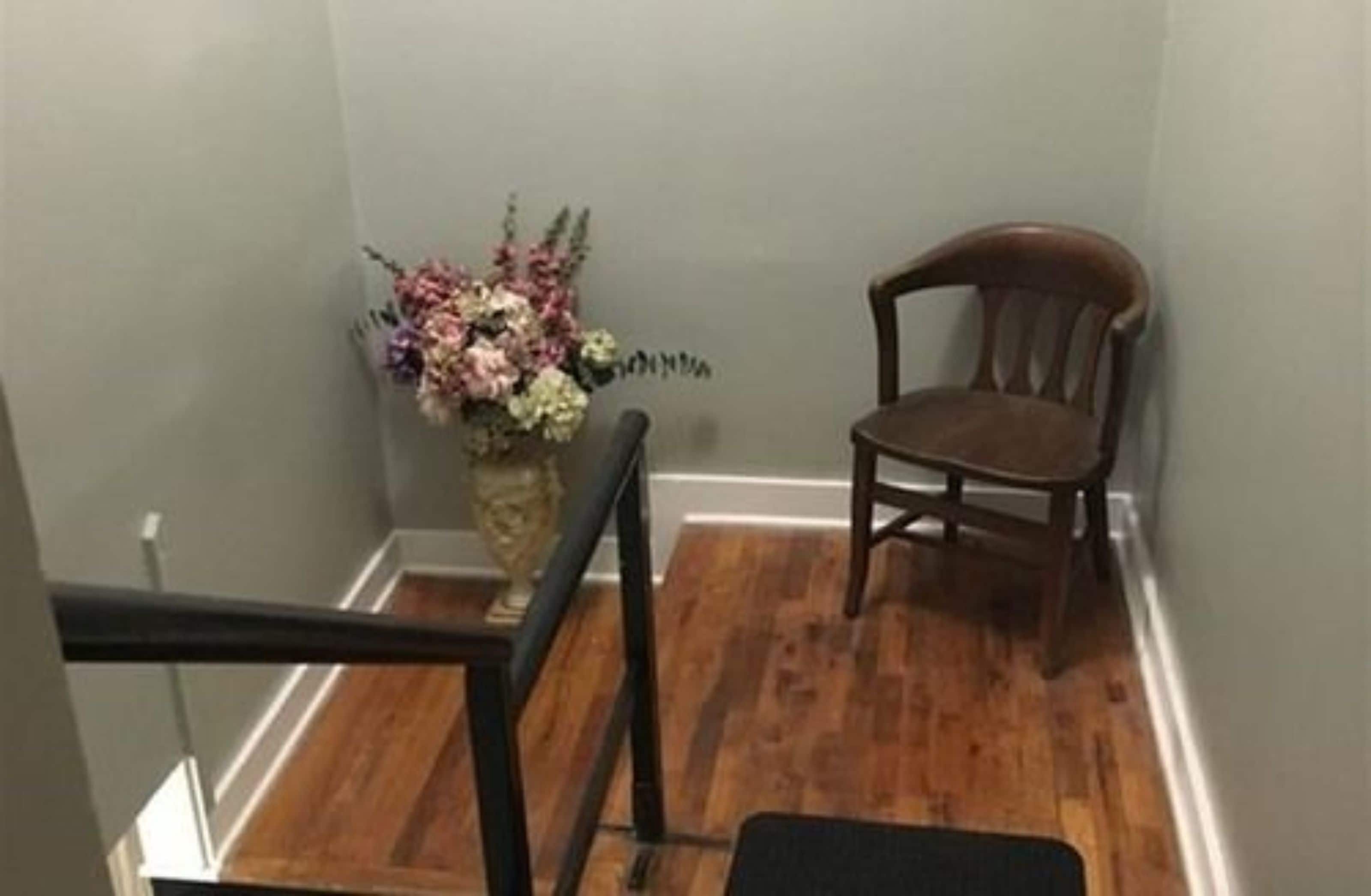 A corner of a room with a wooden floor, a chair, and a vase of flowers.
