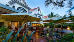 An outdoor dining area with multiple tables and umbrellas, surrounded by lush greenery.