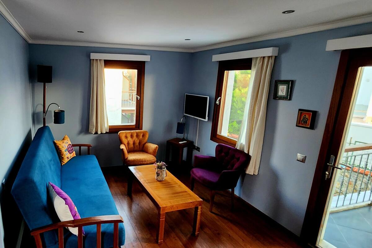 A compact room with a sofa, a wooden table, a purple armchair, and a TV on the wall.
