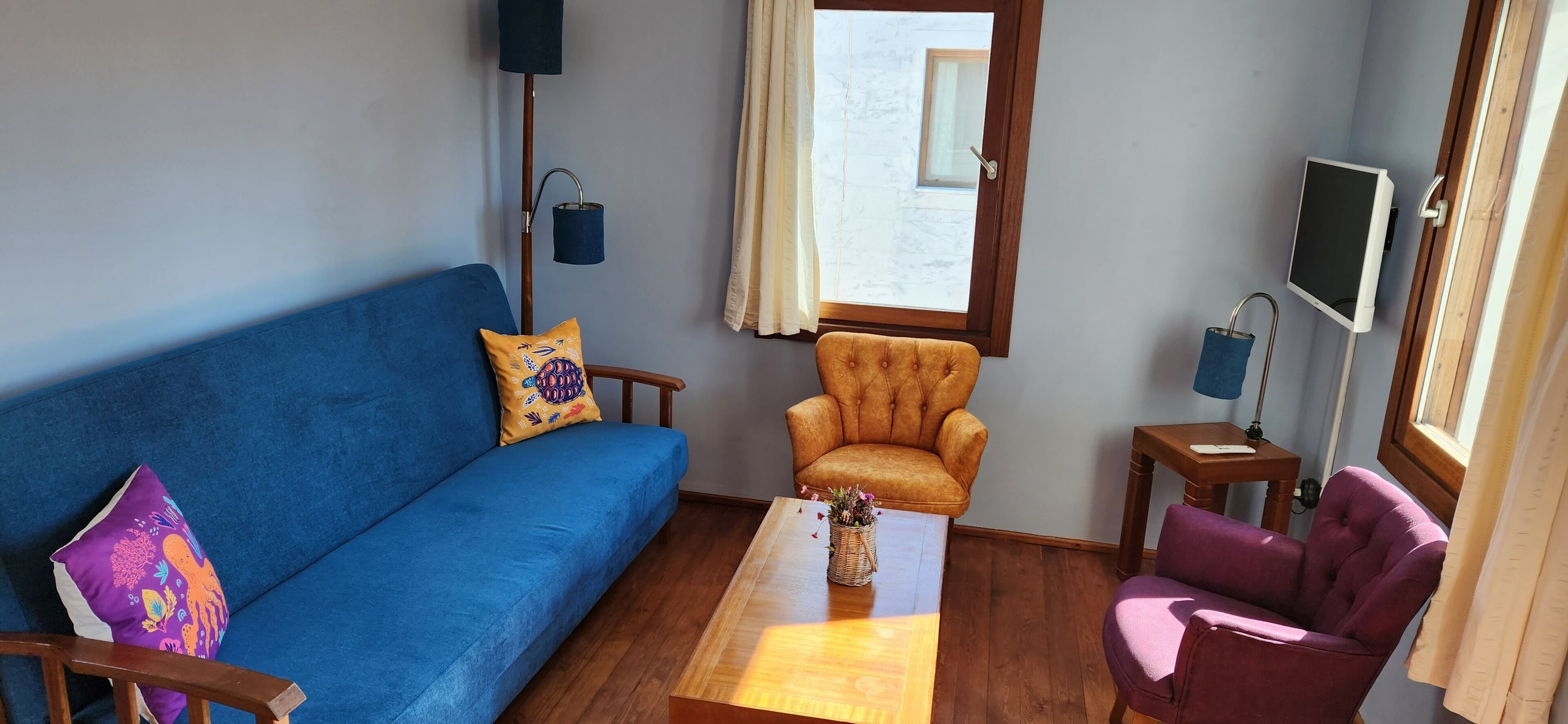 Una sala de estar con un sofá azul, una mesa de centro de madera, un sillón amarillo y un sillón morado, y un televisor sobre un mueble.