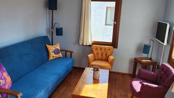 Una sala de estar con un sofá azul, una mesa de centro de madera, un sillón amarillo y un sillón morado, y un televisor sobre un mueble.