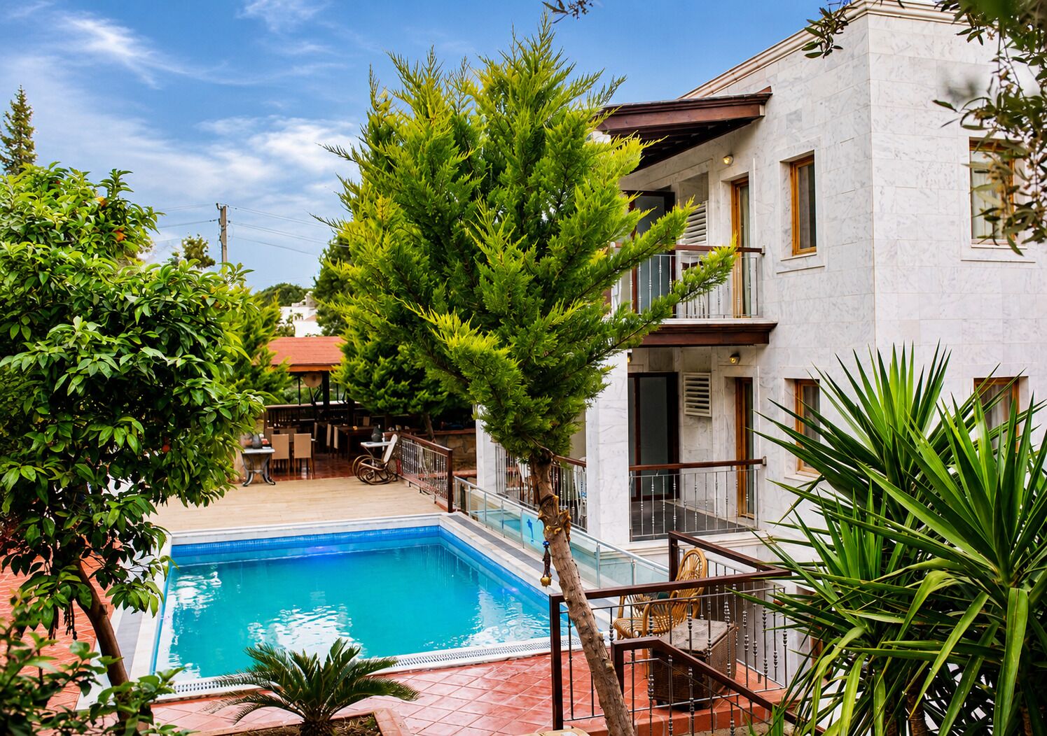 A swimming pool with a wooden deck, surrounded by lush greenery and a two-story building with balconies.
