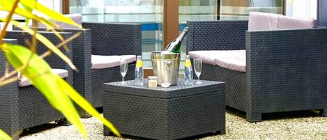 An outdoor seating area with wicker furniture, a table with a bottle and glasses, and a glass door in the background.