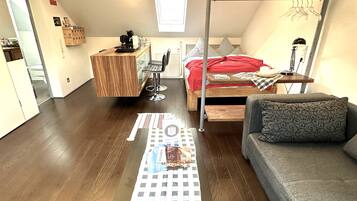Ein kleiner, gut beleuchteter Raum mit Holzboden, einem Bett mit roter Decke, einer Couch, einem Tisch mit kariertem Tischläufer und einem Stockbett mit rotem Kopfkissen.