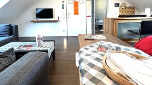 A modern loft apartment with a high ceiling, exposed wooden beams, a flat-screen TV mounted on the wall, a cozy seating area with a sofa and a small table, and a kitchen area with a microwave and a wooden cabinet. - Schacht85 (Oberhausen)