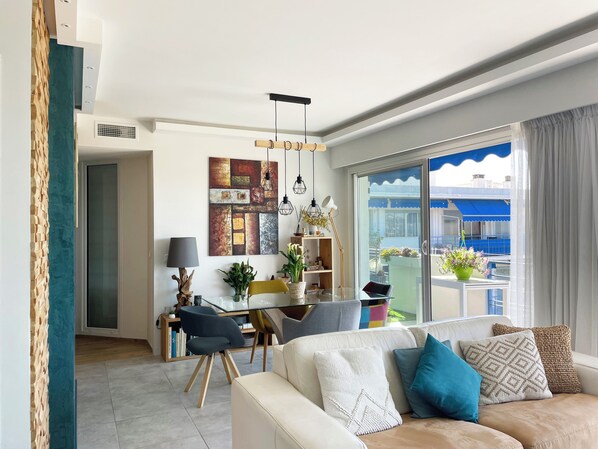 A modern living room with a sofa, a dining area with a table, and a balcony with plants.