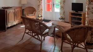 A cozy living room with a wooden table, wicker chairs, a television, and a door with red shutters. - Apartment in a Provencal farmhouse in the Orange countryside (Orange)