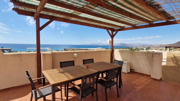 Une terrasse avec une table et des chaises en bois, un ventilateur de plafond et une vue sur la mer et les montagnes.